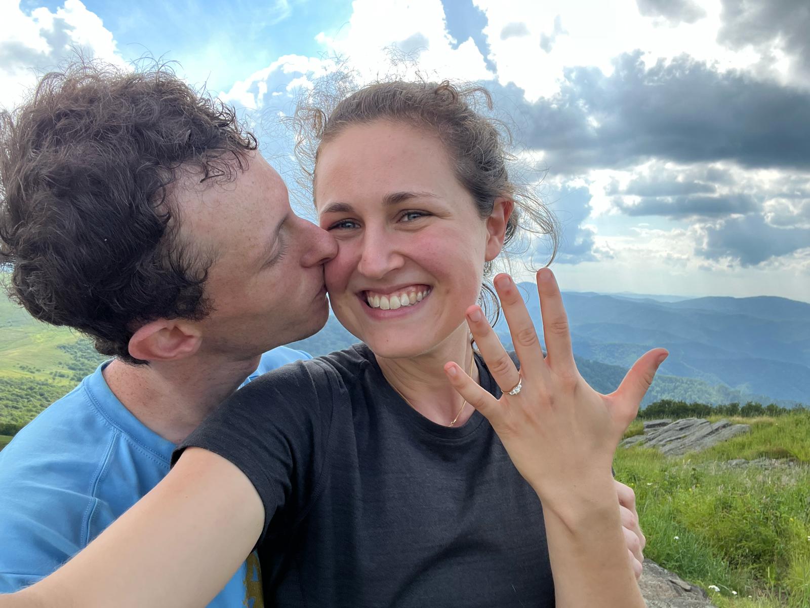 Oren kisses Ella’s cheek while she shows her engagement ring, mountains behind